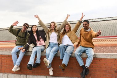 Beş neşeli öğrenci, alçak bir duvarda otururken kollarını kaldırıp gülümsüyor.