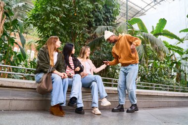 Genç adam botanik bahçesindeki bankta oturan üç kız arkadaşını selamlıyor, konuşuyor ve eğleniyor.