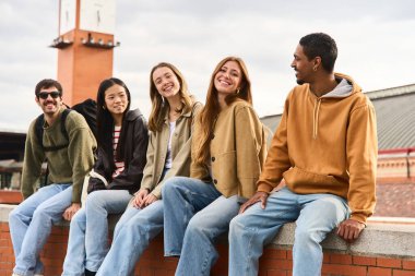 Beş mutlu genç öğrenci tuğla duvarda oturuyor, konuşuyor ve gülümsüyor.