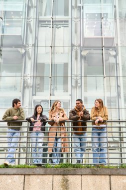 Beş genç öğrenci modern bir binanın balkonunda mutlu bir şekilde sohbet ediyor, molalarının tadını çıkarıyor ve canlı bir şehir manzarasına karşı kahkahalarını paylaşıyorlar.
