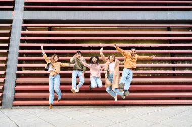 Altı neşeli çoklu etnik kökenli genç öğrenci modern bir binanın önüne atlayıp mutluluklarını ve dostluklarını ifade ediyorlar.