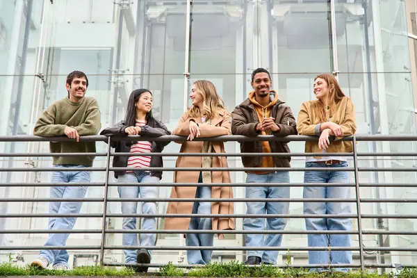 Çok ırklı beş genç, boş zamanlarını modern cam bir binanın önündeki korkuluklara yaslanarak sohbet ederek ve gülümseyerek geçiriyorlar..
