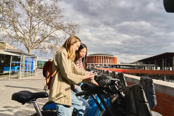 Telefon uygulaması kullanan iki genç kadın, tren istasyonunun yakınındaki bir benzin istasyonunda elektrikli bisiklet kiralıyor.