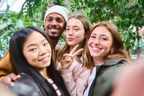 Dört neşeli çoklu etnik genç öğrenci bir serada selfie çekiyorlar, arkadaşlık ve çeşitliliği kutluyorlar.