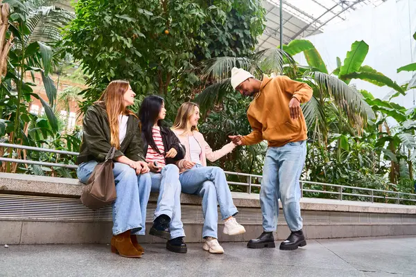 Genç adam botanik bahçesindeki bankta oturan üç kız arkadaşını selamlıyor, konuşuyor ve eğleniyor.