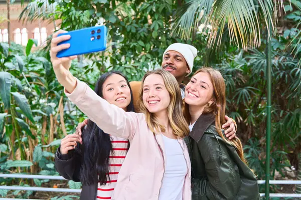 Dört neşeli çoklu etnik öğrenci botanik bahçesindeki bir serada akıllı bir telefonla selfie çekiyor ve birlikte vakit geçiriyorlar.