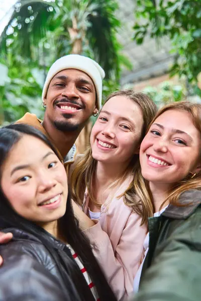Dört neşeli çoklu etnik üniversite öğrencisi kampüs serasında selfie çektirip birlikte vakit geçiriyorlar.