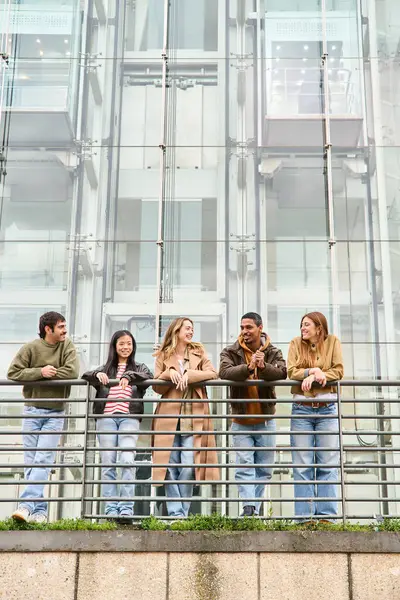 Beş genç öğrenci modern bir binanın balkonunda mutlu bir şekilde sohbet ediyor, molalarının tadını çıkarıyor ve canlı bir şehir manzarasına karşı kahkahalarını paylaşıyorlar.