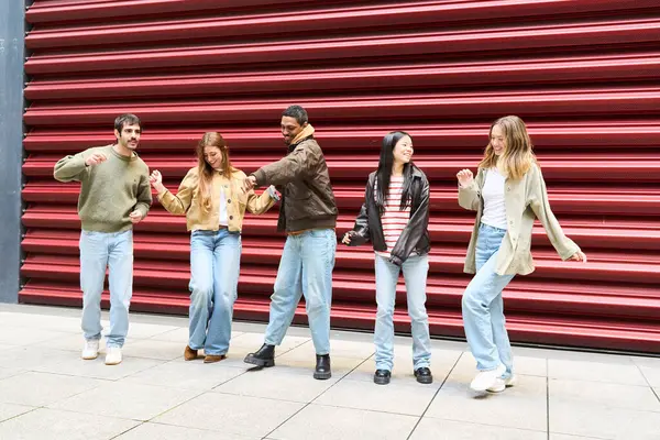 Beş çok ırklı arkadaş kırmızı metalik bir duvarın önünde el ele tutuşup dans ediyorlar.