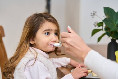 Annem yemek masasında otururken komik surat yapan küçük Latin kızına yoğurt yediriyor.