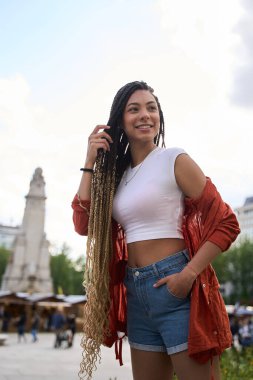 Uzun örgülü, beyaz bluzlu ve kot şortlu gülümseyen genç kadın, şehir meydanında güneşli bir günün tadını çıkarıyor.