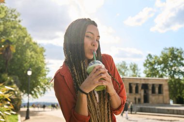 Parkta güneşli bir günde soğuk bir içecek içen uzun örgülü genç bir kadın. Arka planda dekort tapınağı var.