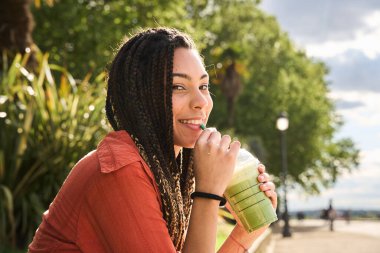 Uzun örgülü, mutlu genç bir kadın yaz günü parkta serinletici bir buzlu yeşil çay içiyor.