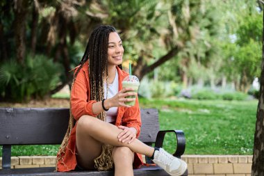 Uzun örgülü genç bir kadın güneşli bir yaz gününde bankta oturmuş soğuk bir içeceğin tadını çıkarıyor.