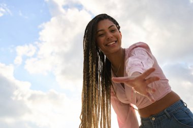 Uzun örgülü, gülümseyen genç bir kadın elini bulutlara ve mavi gökyüzüne uzatıyor.