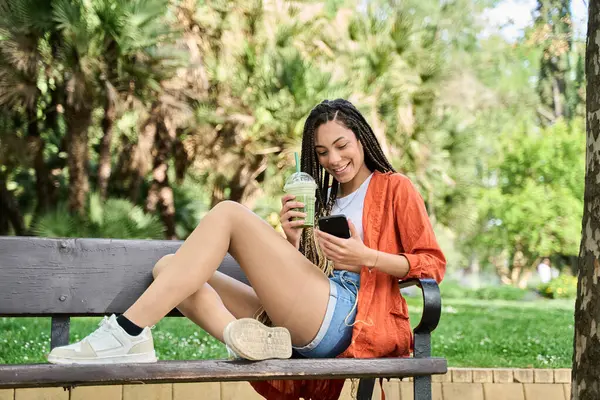 Saçları örgülü mutlu genç bir kadın ferahlatıcı bir içki içiyor ve parkta bankta otururken telefonunu kullanıyor.