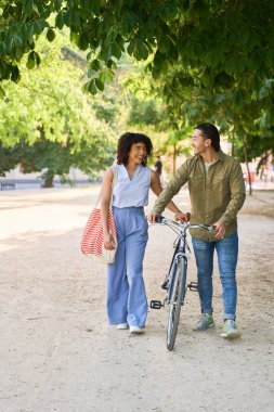 Bisikletle parkta yürüyen, sohbet ve yaz havasının tadını çıkaran çok ırklı genç bir çift.
