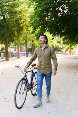 Mutlu bir adam ağaçlık bir parkta klasik bisikletiyle yürüyor, açık havada güneşli bir günün tadını çıkarıyor.