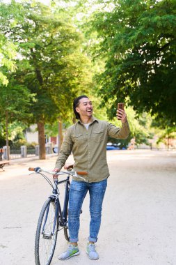 Bisikletiyle parkta yürüyen mutlu genç adam, gün ışığında güneşlenirken akıllı telefonuyla video görüşmesinin tadını çıkarıyor.