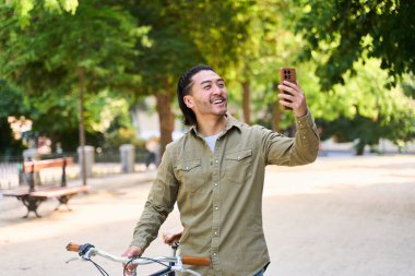 Şehir parkında bisikletiyle yürürken akıllı telefonuyla selfie çeken mutlu adam yaz gününün tadını çıkarıyor.