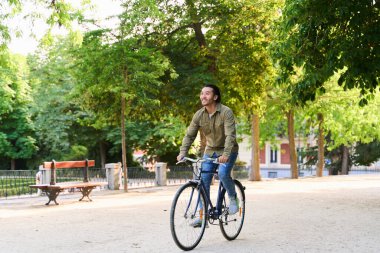 Genç adam canlı yeşil bir şehir parkında bisikletini sürüyor, güneş ışığını emiyor ve dışarıda kaygısız bir yaz gününün tadını çıkarıyor.