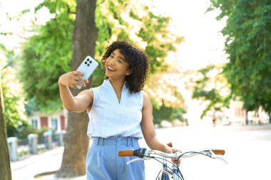 Gülümseyen iş kadını, bisiklet parkında yürürken akıllı telefonuyla selfie çekiyor ve yaz gününün tadını çıkarıyor.