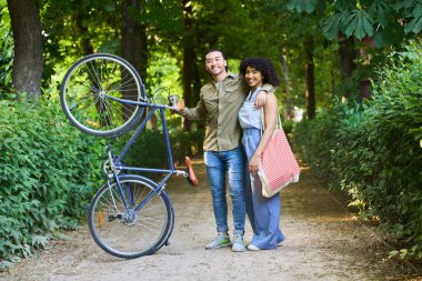 Bisikletleriyle yeşil bir parkta kucaklaşıp poz veren, sürdürülebilir bir yaşam tarzının ve birlikte kaliteli zaman geçirmenin tadını çıkaran çok ırklı genç bir çift.