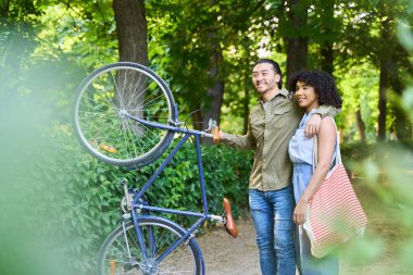 Genç çoklu etnik bir çift parkta bisiklet ve çanta taşırken birlikte sakin bir öğleden sonranın tadını çıkarıyorlar.
