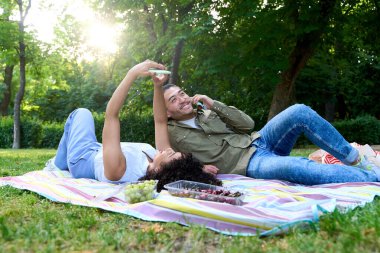 Genç çift battaniyeye uzanmış, parkta pikniğin tadını çıkarıyor, sıcak yaz güneşinin altında selfie çekiyor ve telefonda sohbet ediyorlar.