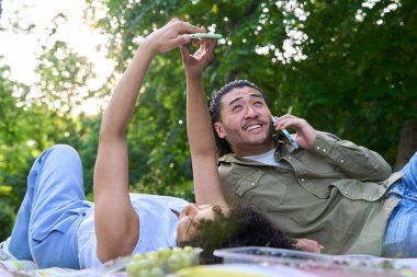 Genç çoklu etnik çift parkta piknik battaniyesinde dinleniyor, selfie çekiyor ve akıllı telefondan konuşuyor.