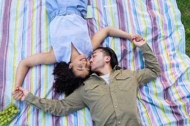 Renkli piknik battaniyesinde uzanan romantik çift, el ele tutuşup öpüşüyor, dışarıda rahatça eğleniyor.