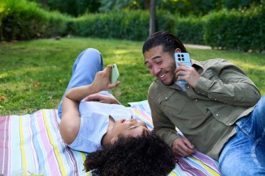 Genç çoklu etnik çift piknik battaniyesinde dinleniyor, birlikte zaman geçiriyorlar ve canlı bir park ortamında akıllı telefonları kullanıyorlar.
