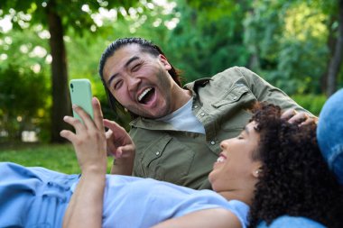 Genç çift birlikte gülüyor ve eğleniyor. Güneşli bir parkta çimlerin üzerinde dinlenirken cep telefonu kullanıyorlar.