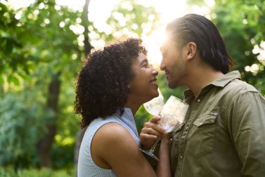 Romantik çift şarap kadehlerini paylaşıyor, gülümseyerek birbirlerine bakıyor, gün batımında doğayla iç içe geçen bir sevgi anının tadını çıkarıyorlar.
