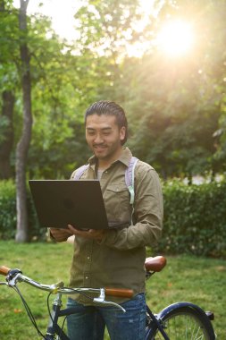 Asyalı serbest çalışan gülümseyen ve yanında bisikletiyle parkta dizüstü bilgisayarıyla çalışan, gün ışığının keyfini çıkaran