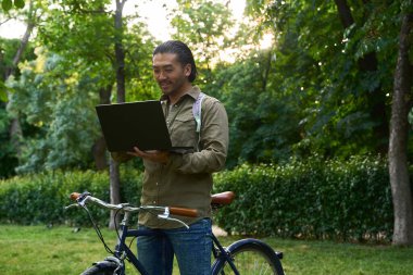 Bisikletiyle parkta dizüstü bilgisayarda çalışan Asyalı serbest yazar temiz hava ve güneş ışığının tadını çıkarıyor.