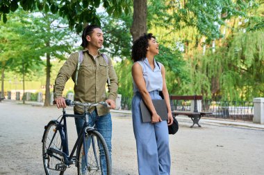 Yeşil bir parkta yürüyen ve gülen iki genç profesyonel, biri bisikleti itiyor ve diğeri de dizüstü bilgisayar taşıyor.