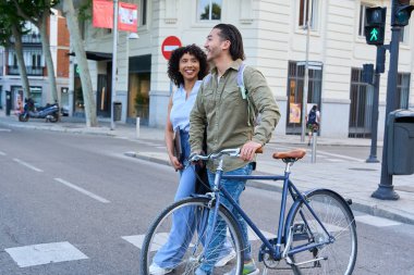 İki genç profesyonel yaya geçidinde karşıdan karşıya geçiyor, bisikletinin yanında yürüyor, ikisi de gülümsüyor ve başka tarafa bakıyor.