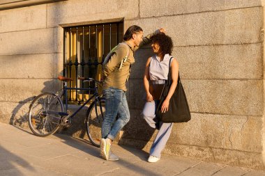 Genç bir adam ve kadın güneşli bir caddede sohbet ediyorlar, bir bisiklet duvara yaslanmış, neşe ve bağlantı yayıyorlar.