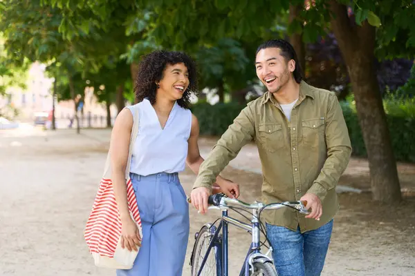 Genç, çok ırklı bir çift parkta yürürken ve gülerken antika bir bisikleti itiyorlar.