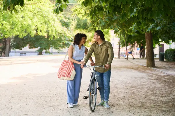 Genç bir çift parkta birlikte yürüyor, romantik bir yürüyüşün keyfini çıkarırken adam yanlarında bisikleti itiyor.