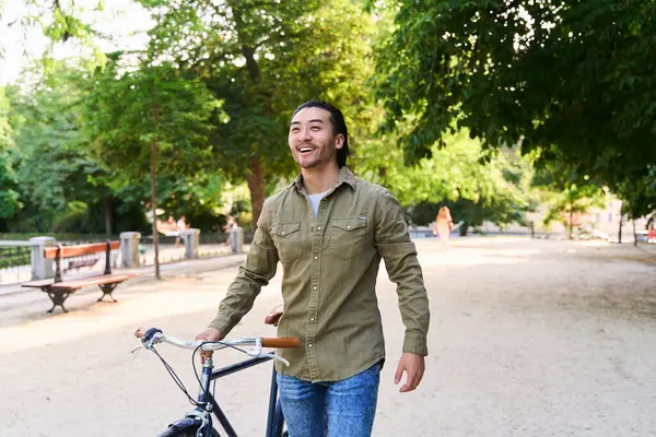 Neşeli bir şehir parkında bisikletiyle yürüyüp güneşin tadını çıkaran ve dışarıda keyifli bir gün geçiren mutlu genç adam.