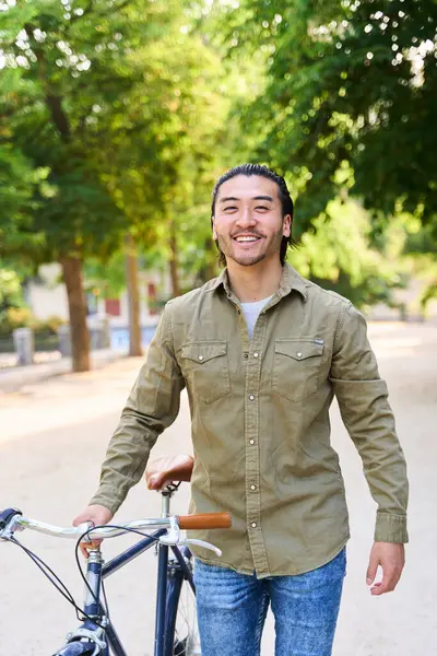 Asyalı genç bir adam, güzel bir günde güneşin tadını çıkaran ağaçlık bir parkta, eski bisikletiyle birlikte yürüyor.