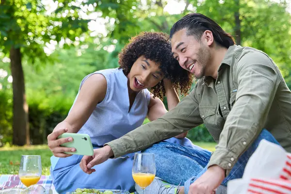 Parkta romantik bir pikniğin tadını çıkaran mutlu çoklu etnik çift. Güneşli gökyüzünün altında akıllı telefonlarıyla gülüp selfie çekiyorlar.