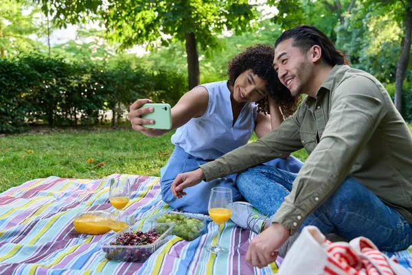 Genç çoklu etnik çift parkta romantik bir pikniğin tadını çıkarıyor, gülümsüyor ve akıllı telefonlu bir özçekim ile neşeli anları yakalıyor.