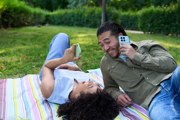 Genç çoklu etnik çift piknik battaniyesinde dinleniyor, birlikte zaman geçiriyorlar ve canlı bir park ortamında akıllı telefonları kullanıyorlar.