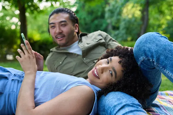 Mutlu çoklu etnik çiftler güneşli bir parkta battaniyenin üzerinde dinleniyorlar, akıllı telefonlarıyla gülüyorlar ve birbirlerinin arkadaşlığından keyif alıyorlar.