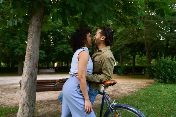 Genç çok ırklı çift parkta romantik bir anı paylaşıyor, yemyeşil ve güneş ışığıyla çevrili bir bisikletin yanında neşeyle kucaklaşıyorlar.