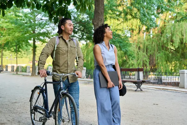 Yeşil bir parkta yürüyen ve gülen iki genç profesyonel, biri bisikleti itiyor ve diğeri de dizüstü bilgisayar taşıyor.