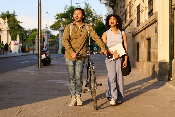 İki mutlu öğrenci kaldırımda yürüyorlar, güneşli bir günün tadını çıkarıyorlar, dizüstü bilgisayar taşıyorlar ve bisiklet sürüyorlar.